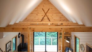 A wooden cabin with a sloped roof, a black wood-burning stove, and a large window. - The Pines Cabin | New Tiny Home Near Mt Snow! (Wilmington)