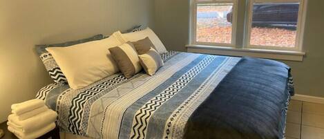 Un dormitorio con cama, almohadas, una mesita de noche con toallas y una ventana con persianas.