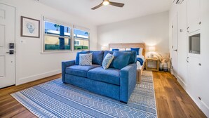 A room with a blue sofa, a wooden headboard, a ceiling fan, and a view of palm trees through a window.
