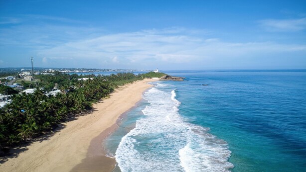 A beach with a long stretch of sand, a lighthouse, and a clear blue sky.
