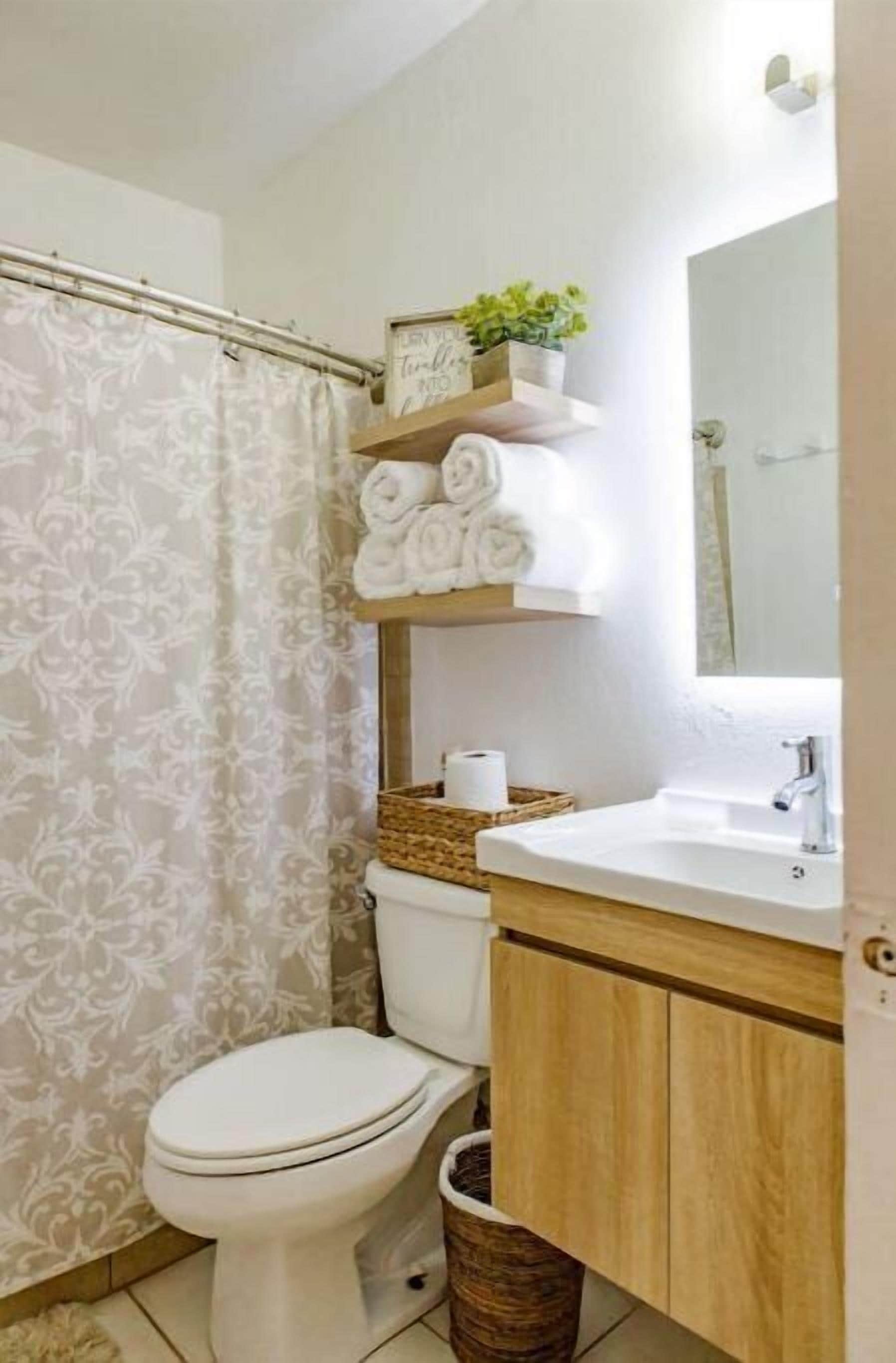 Ein Badezimmer mit Toilette, Waschbecken, Spiegel, Duschvorhang und Ablage mit Handtüchern und Pflanze.