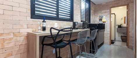 A kitchen with a brick wall, a window with blinds, a bar counter with stools, and a microwave.