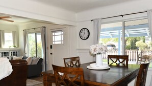 A dining area with a wooden table, chairs, a clock on the wall, and a view through large windows. - Cozy Blue Cottage

 (Willcox)