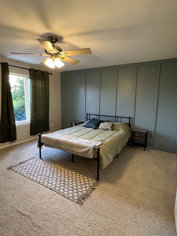 A bedroom with a bed, a ceiling fan, a window with curtains, and a patterned rug. - Green Jay House (Centennial)