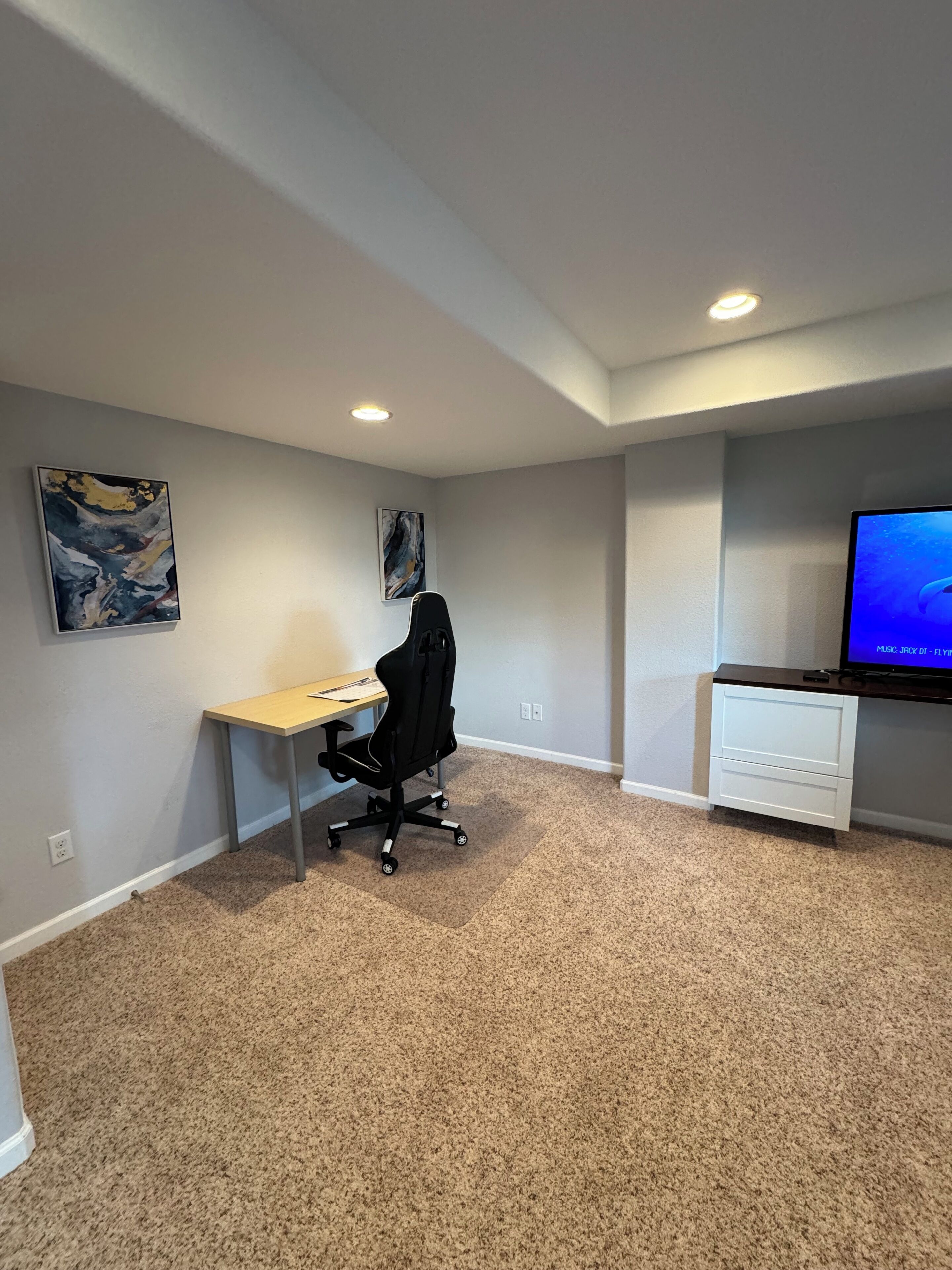 A room with a desk, office chair, two abstract paintings, a television, and a carpeted floor.