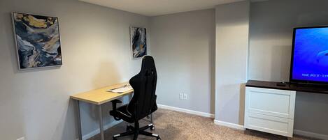 A room with a desk, office chair, two abstract paintings, a television, and a carpeted floor.