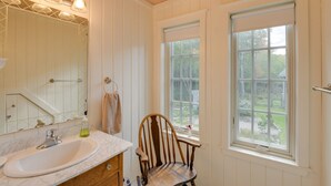 Un baño con lavamanos blanco, espejo y una silla de madera cerca de dos ventanas.