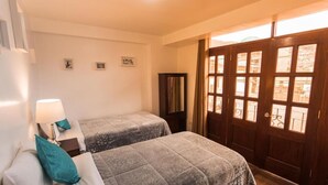 Habitación de hotel con dos camas, una mesita de noche con lámpara, un armario y un ventanal con vistas al exterior.