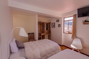 A bedroom with two beds, a wooden wardrobe, a TV mounted on the wall, and a window with a view of buildings.