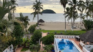 Zona de piscina con vistas a la playa, palmeras y una estructura de techo de paja.