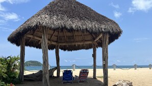 Una cabaña en la playa con techo de paja, dos sillas de playa y vistas al mar.