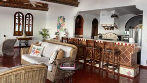 Amplia sala de estar con techo alto, muebles de mimbre, cocina con electrodomésticos modernos y detalles decorativos como un cuadro de colores y un salpicadero de azulejos con motivos tradicionales.
