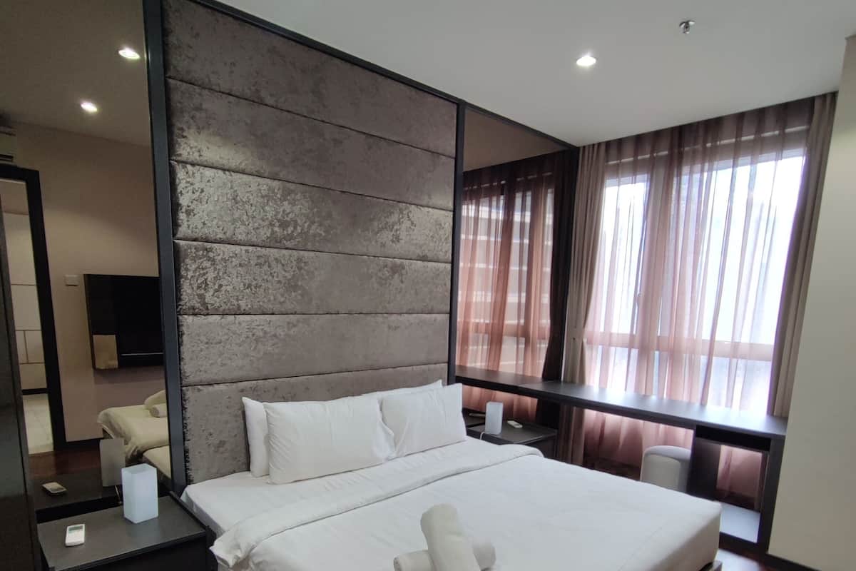 A modern hotel room with a large bed, wooden flooring, a dark headboard, and a large window with curtains.