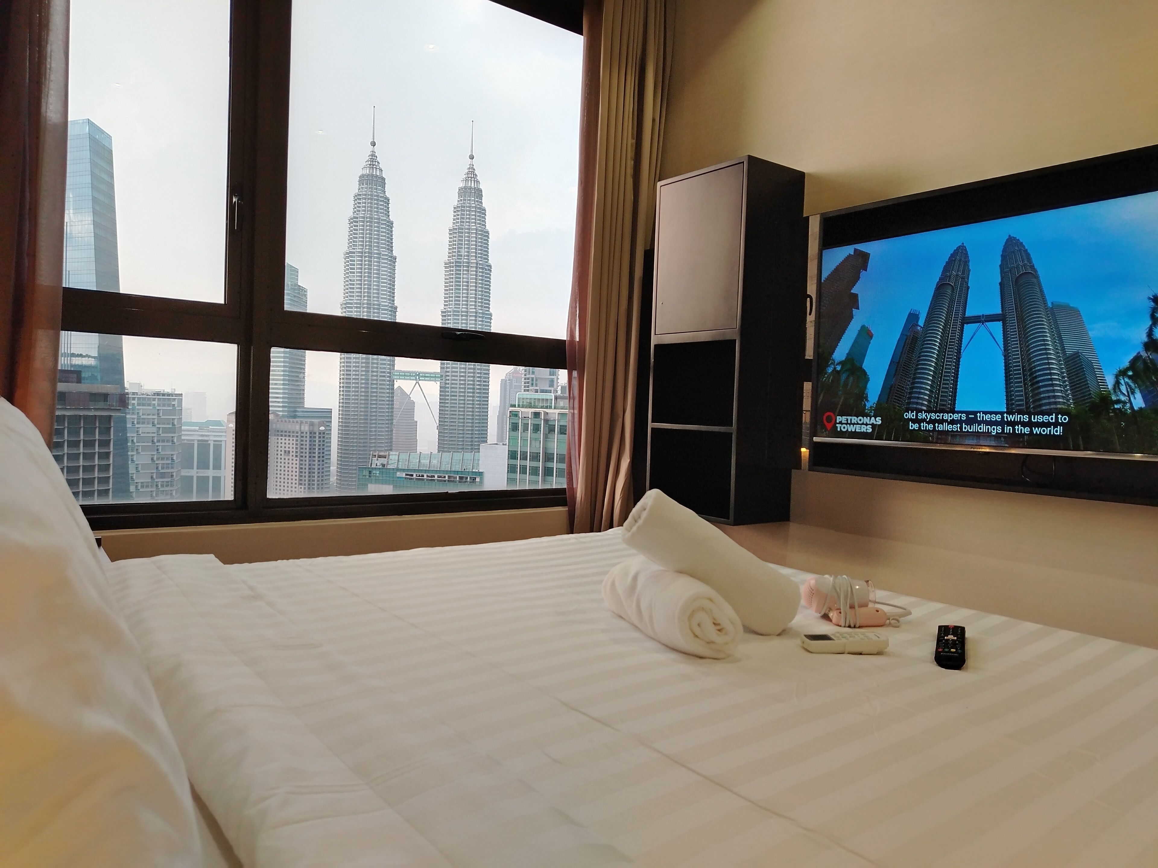A hotel room with a bed, a TV displaying the Petronas Towers, and a view of the cityscape through large windows.