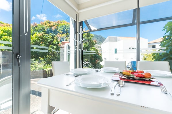 Una mesa de comedor dispuesta con platos, cubiertos y una bandeja de frutas en una habitación luminosa con vistas a zonas verdes y edificios.