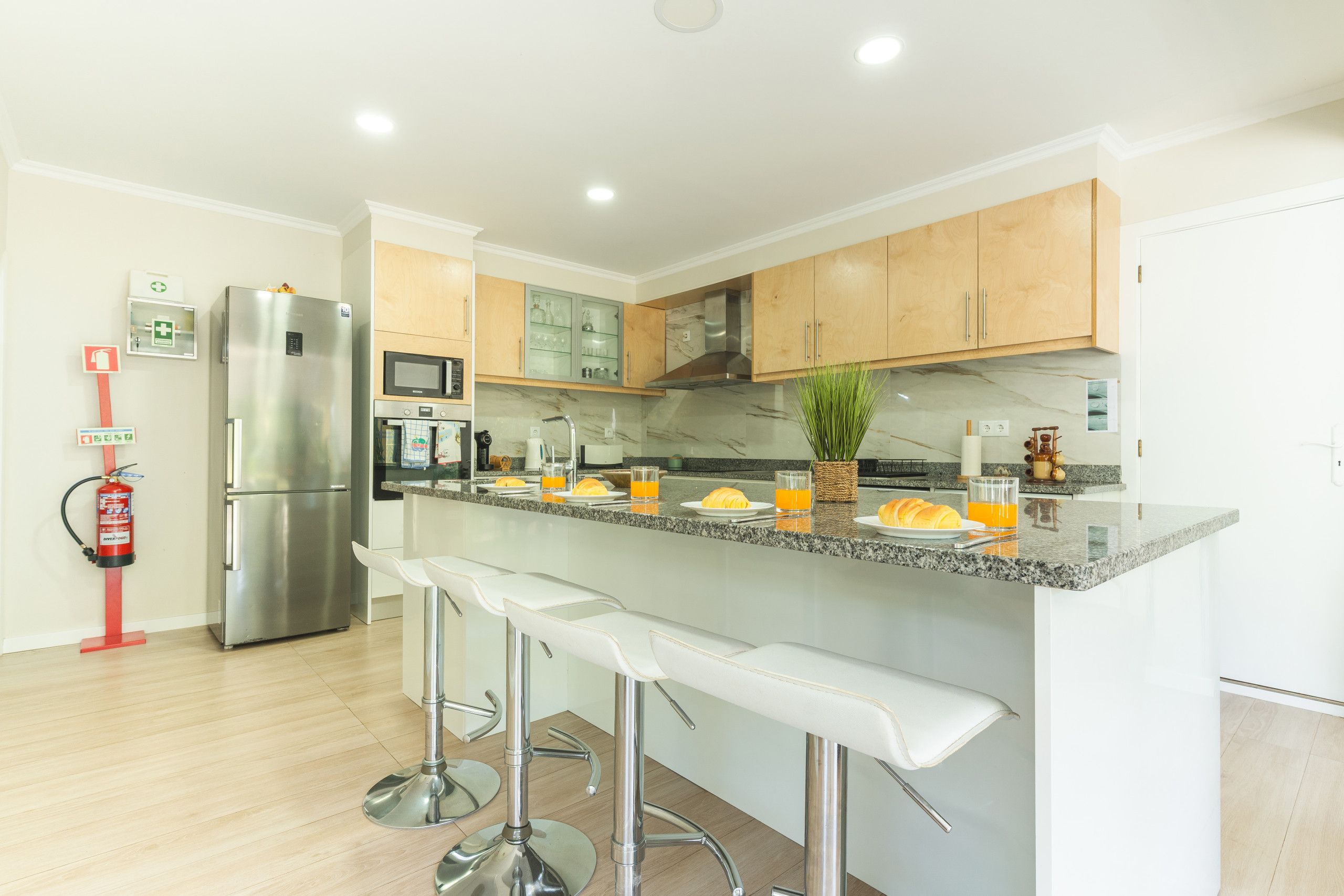 A modern kitchen with a stainless steel refrigerator, a microwave, and a sink. There are bar stools and a plant on the counter.