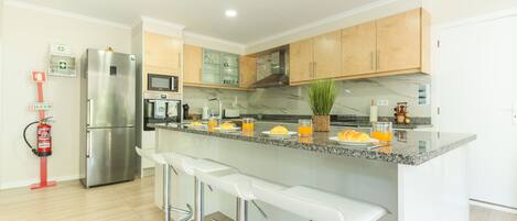 A modern kitchen with a stainless steel refrigerator, a microwave, and a sink. There are bar stools and a plant on the counter.