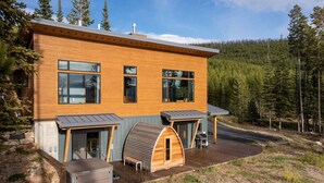 Una casa moderna de madera con ventanas amplias, una zona de estar al aire libre techada y una pequeña construcción de madera.