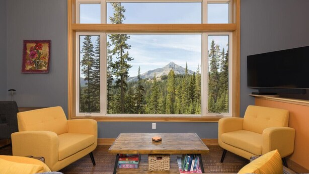 Una sala de estar con dos sofás amarillos, una mesa de centro de madera con libros, un televisor en la pared y un ventanal con vistas a la montaña.