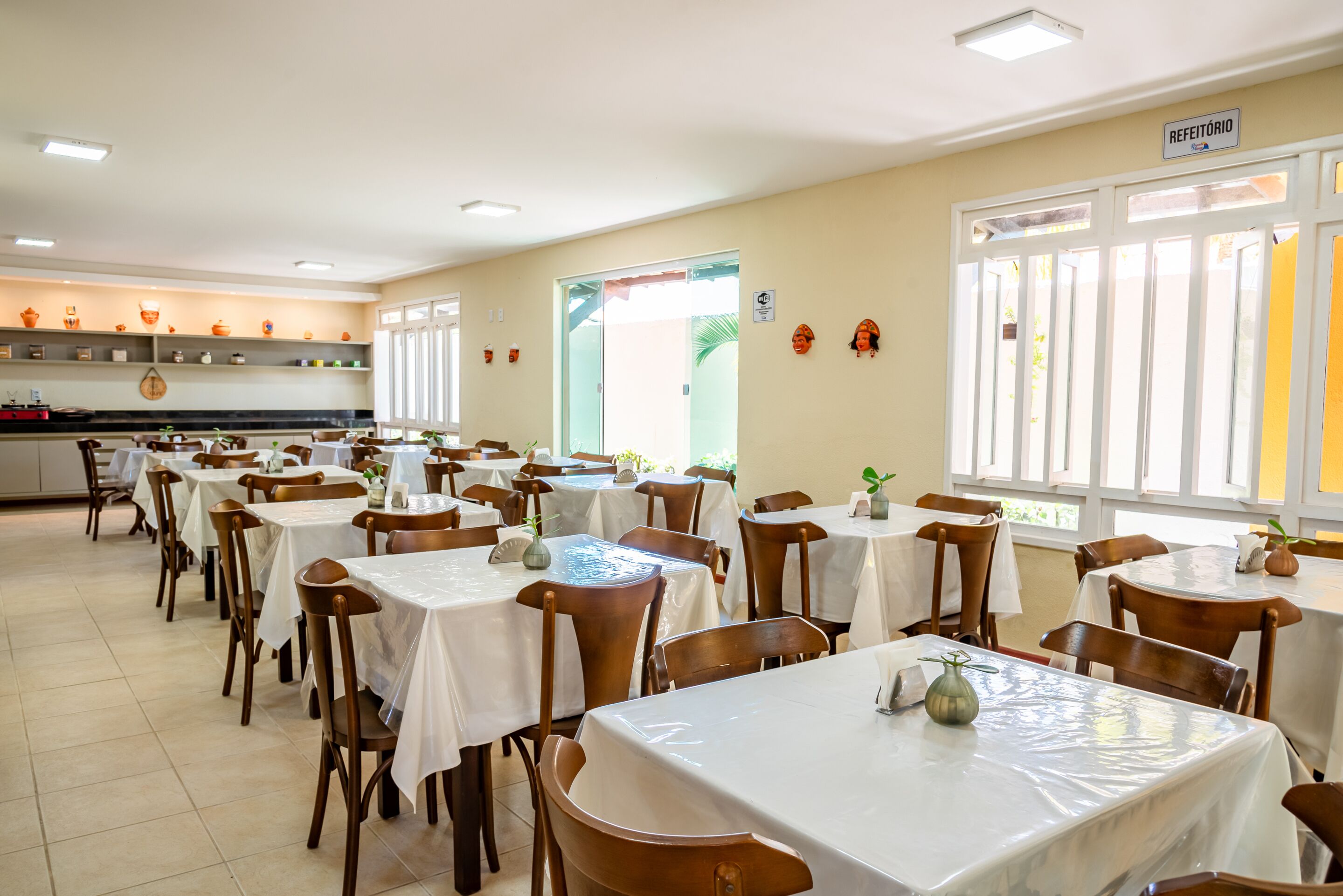 Un comedor con varias mesas y sillas, una cocina al fondo y un cartel que dice "REFEITÓRIO".