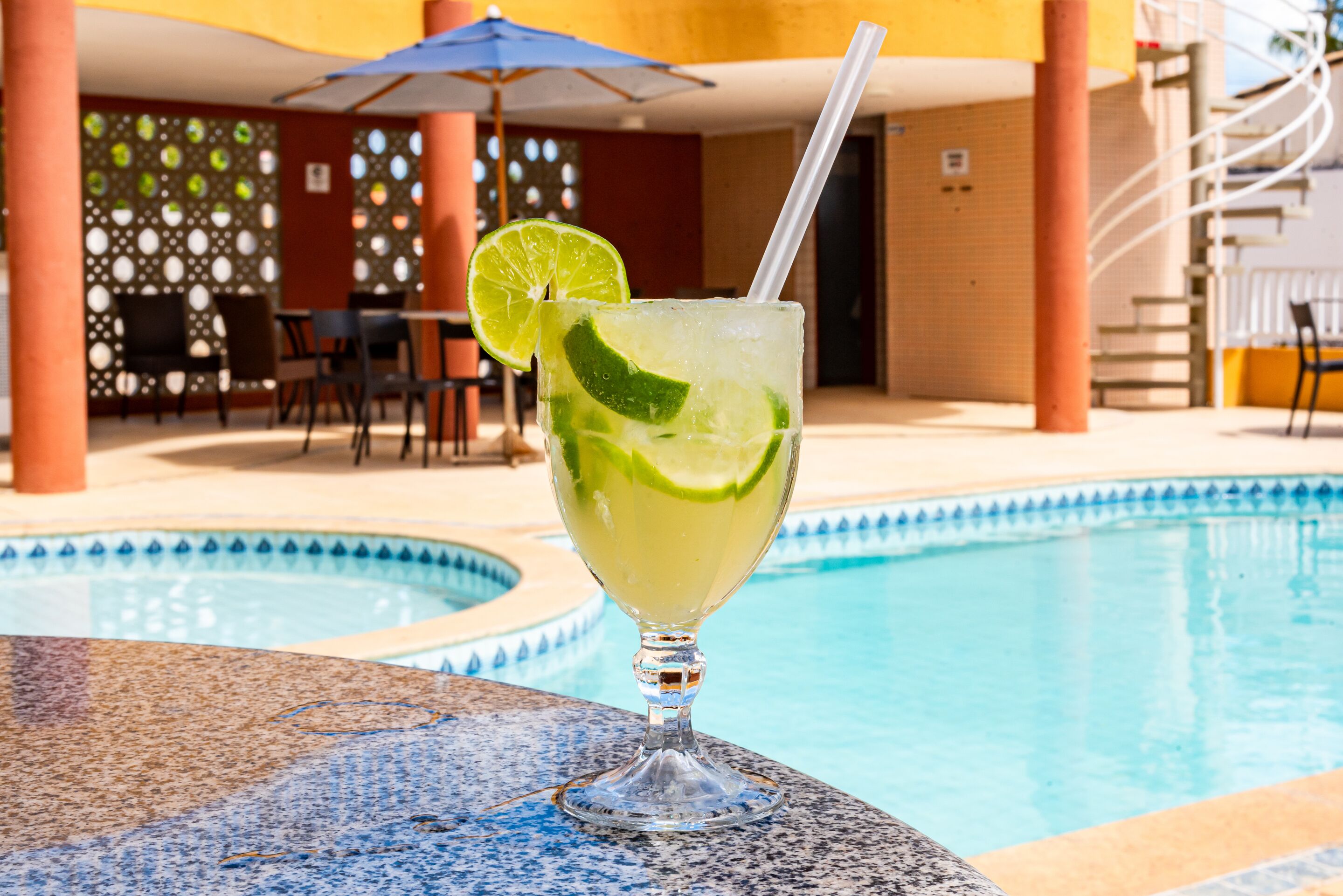 A glass of a lime-based cocktail with a straw, placed on a table by a poolside.