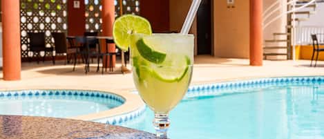 A glass of a lime-based cocktail with a straw, placed on a table by a poolside.