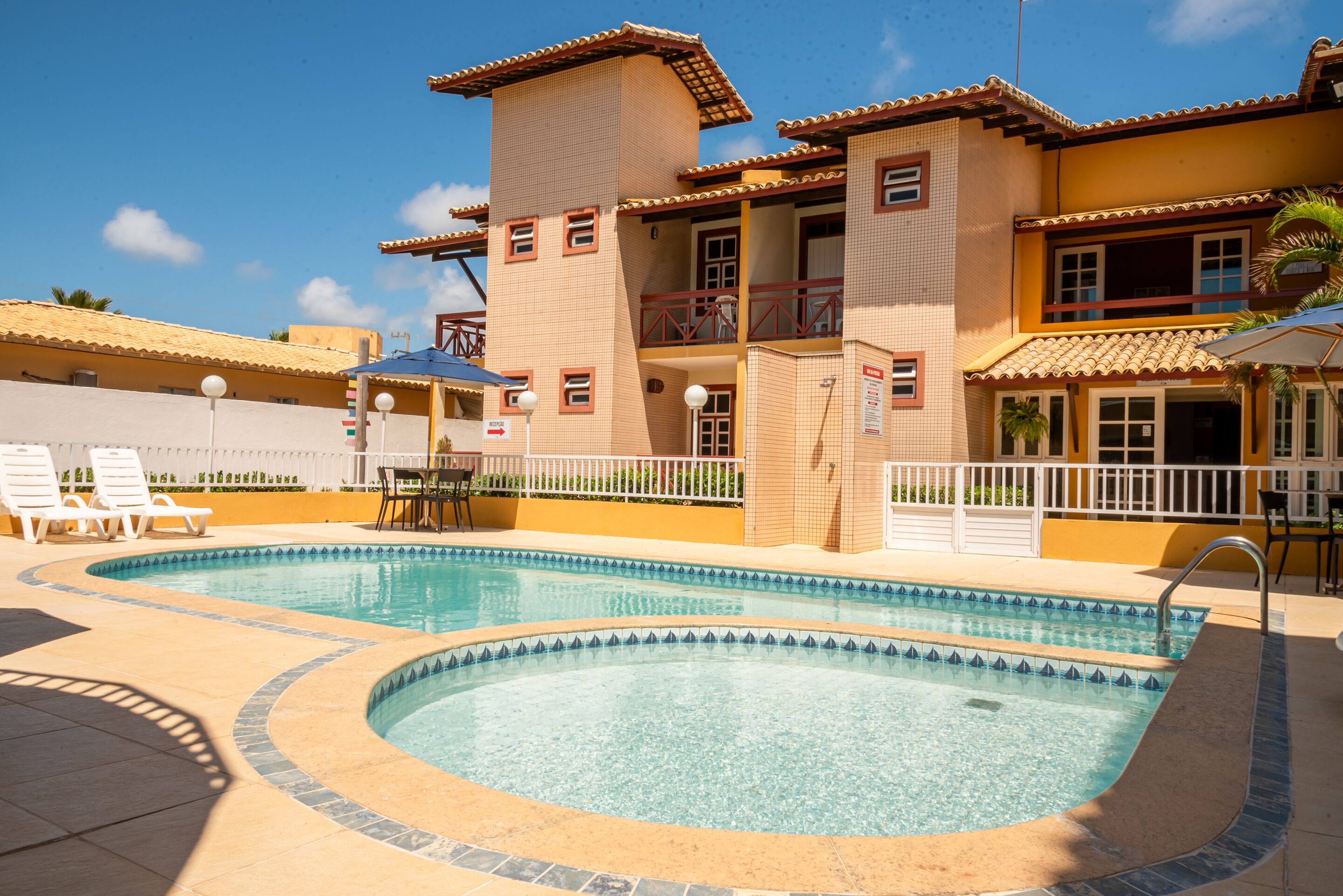 Una piscina con borde de azulejos, rodeada de sillas de descanso y un edificio con tejado y balcones de azulejos.
