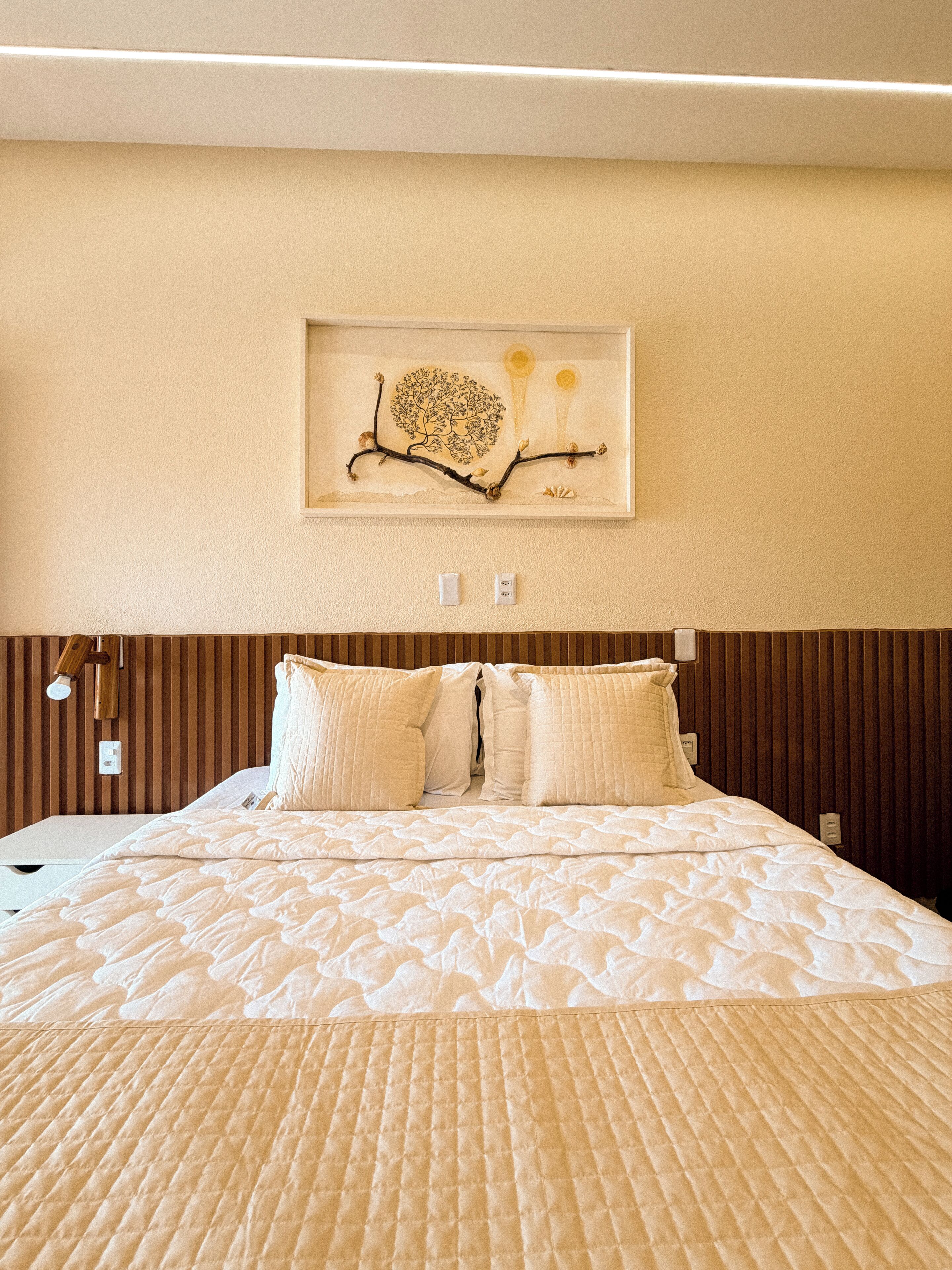 A neatly made bed with a quilted cover, a wall-mounted artwork depicting a tree and sun, and a wooden headboard.