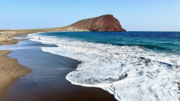 Ein Strand mit einer großen Felsformation, klarem blauen Wasser und einem sandigen Ufer.