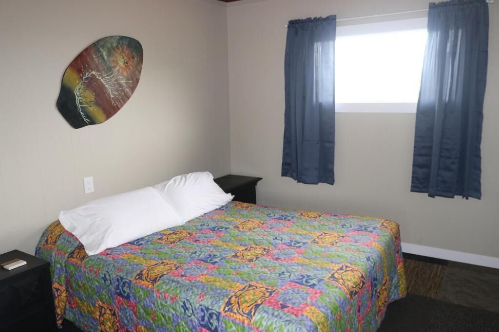 A single bed with a colorful quilt, a nightstand, a wall-mounted painting, and a window with curtains.