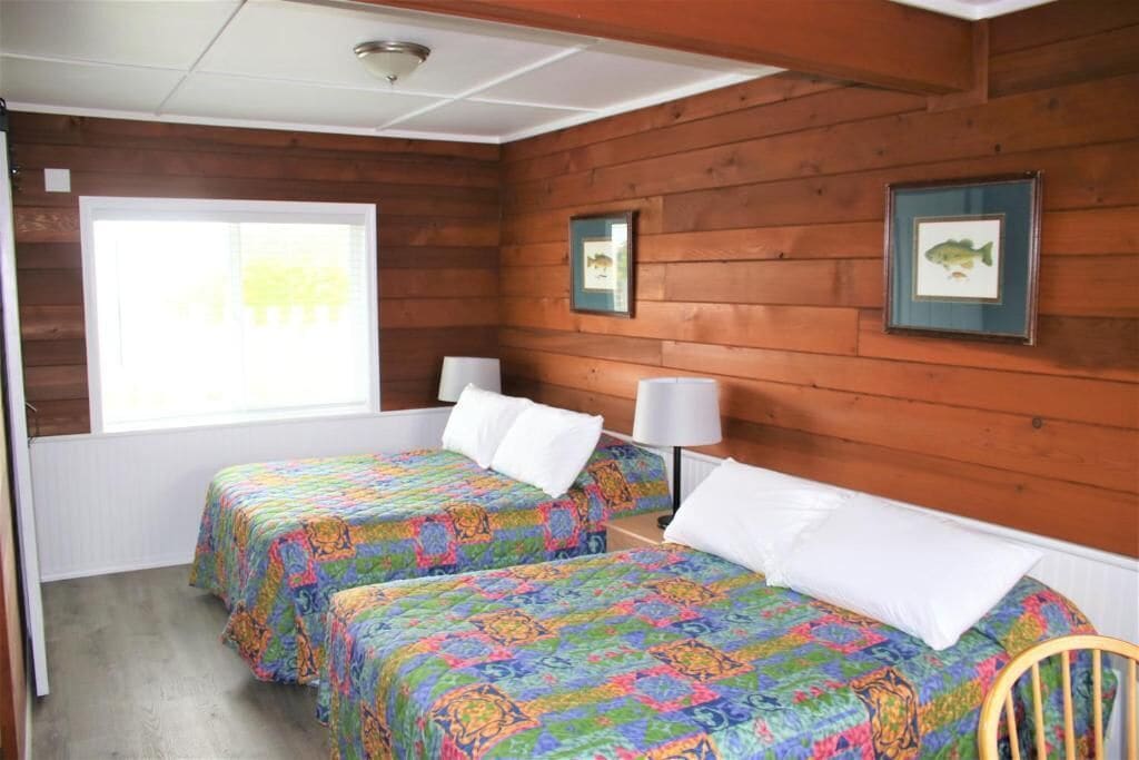 A room with two beds, wooden walls, framed pictures, and a window.