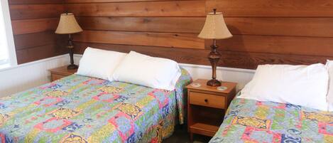 A room with two beds, each with a colorful quilt, wooden headboard, and a nightstand with a lamp.