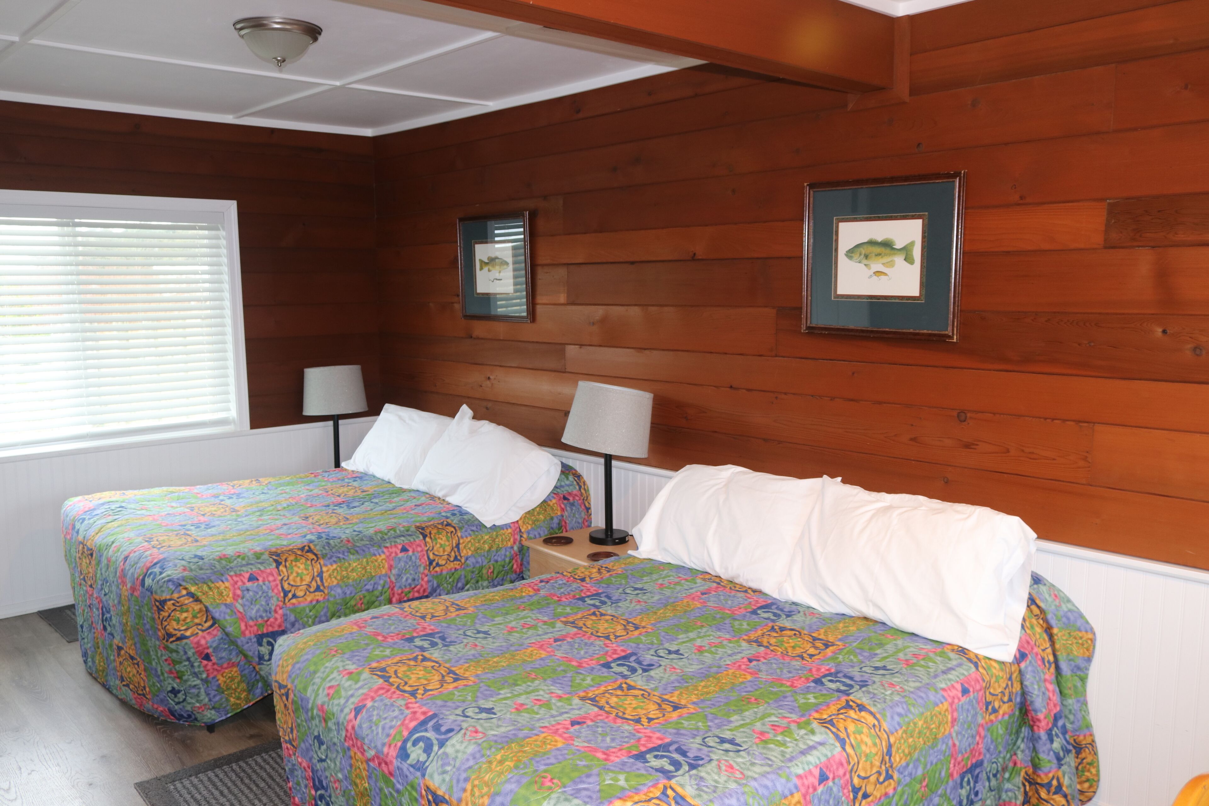 A room with two beds, wooden walls, and framed pictures on the wall.