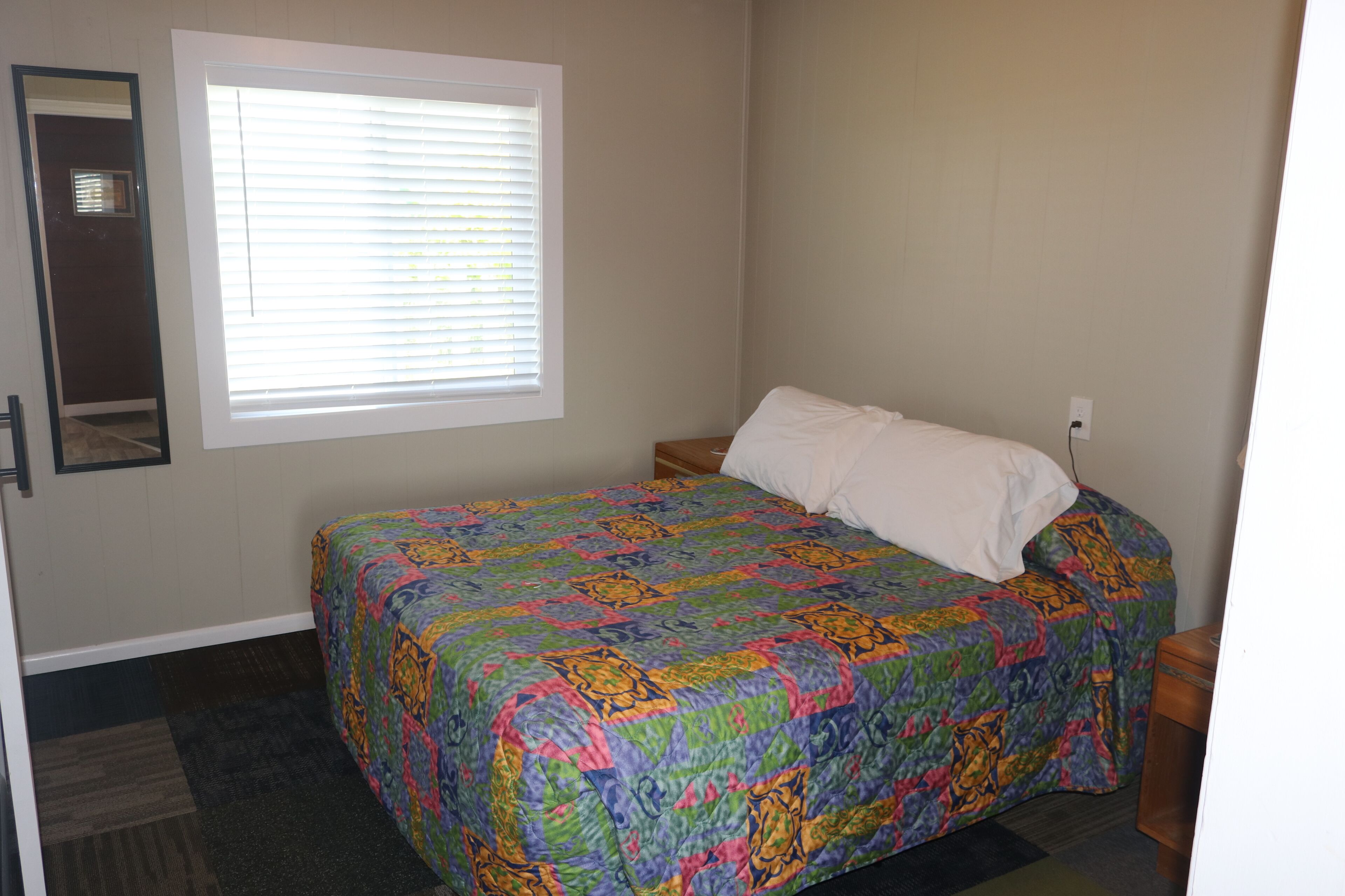 A bedroom with a bed, a window with blinds, a nightstand, and a wall-mounted mirror.