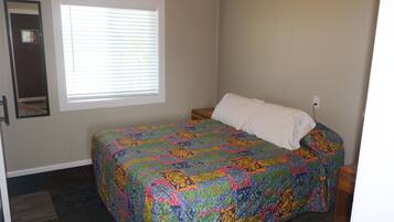 A bedroom with a bed, a window with blinds, a nightstand, and a wall-mounted mirror.