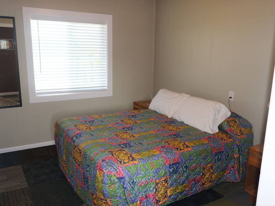 A bedroom with a bed, a window with blinds, a nightstand, and a wall-mounted mirror.