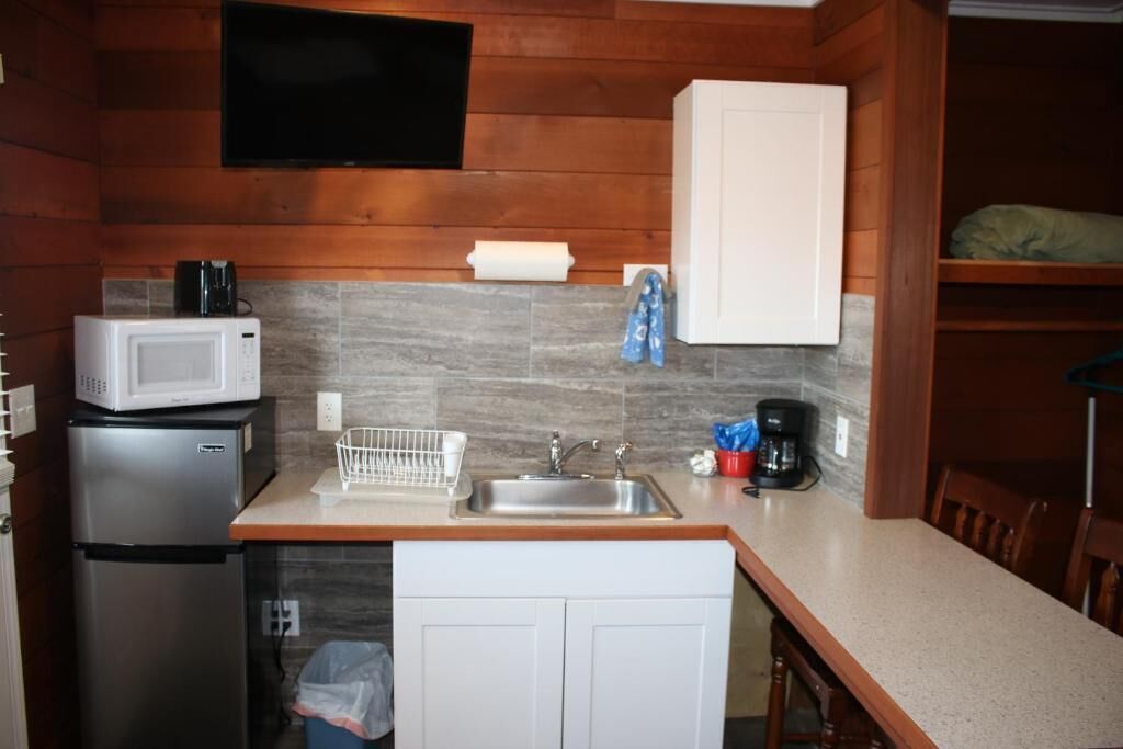 A compact kitchen with a microwave, refrigerator, sink, and a flat-screen TV mounted on the wall.