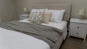 A neatly made bed with a grey blanket and multiple pillows, including a decorative one. There are two bedside tables with lamps and a small drawer.