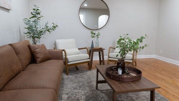 A living room with a brown sofa, two wooden chairs, a coffee table with a woven basket, and two potted plants.