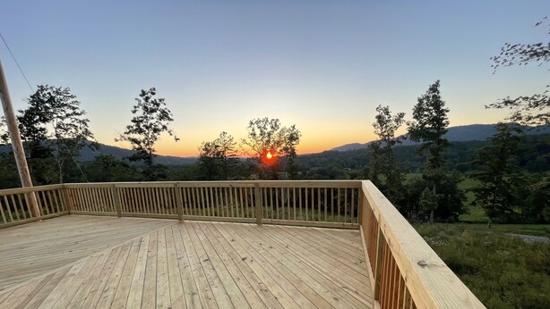 A wooden deck with a railing, overlooking a scenic landscape with trees and a sunset.