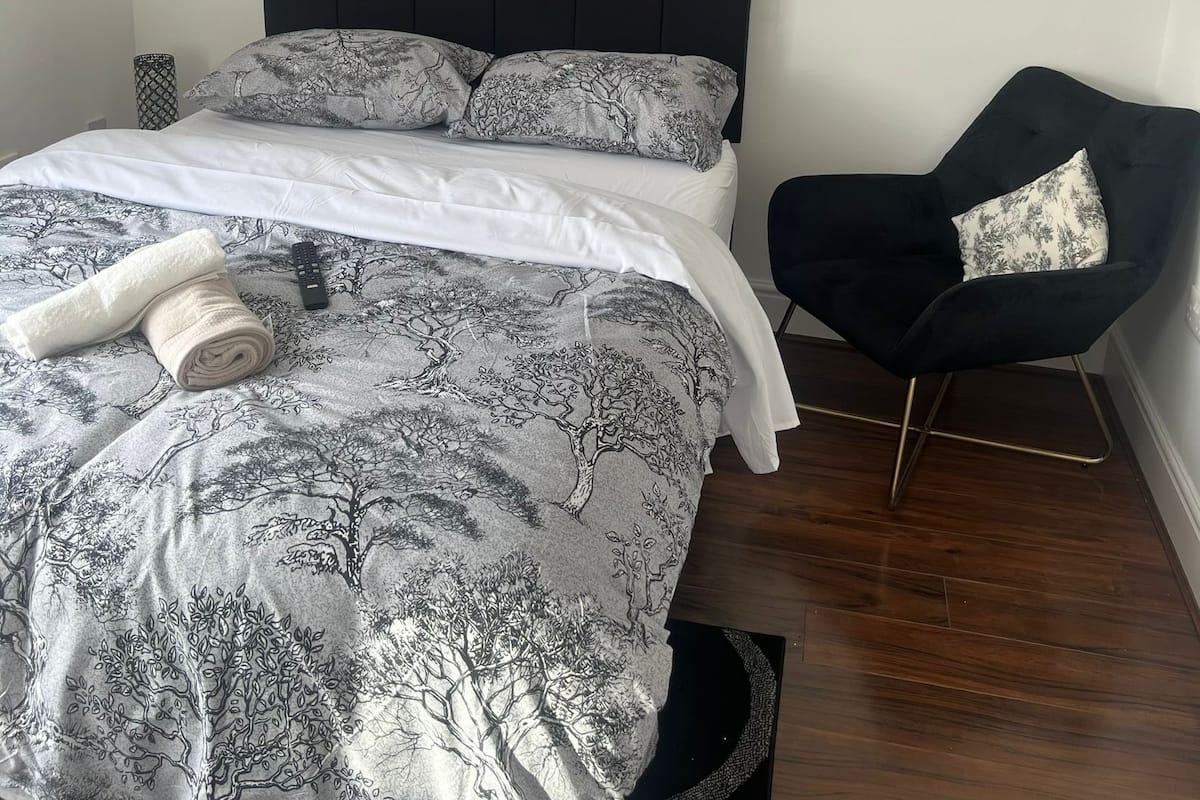A bedroom with a bed, a chair, a framed picture on the wall, and a rolled towel on the bed.