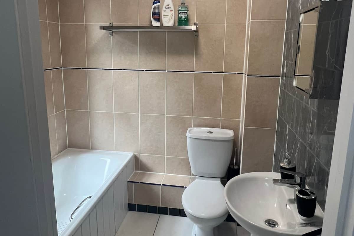 A bathroom with a bathtub, toilet, and sink. There is a shelf with toiletries above the toilet.