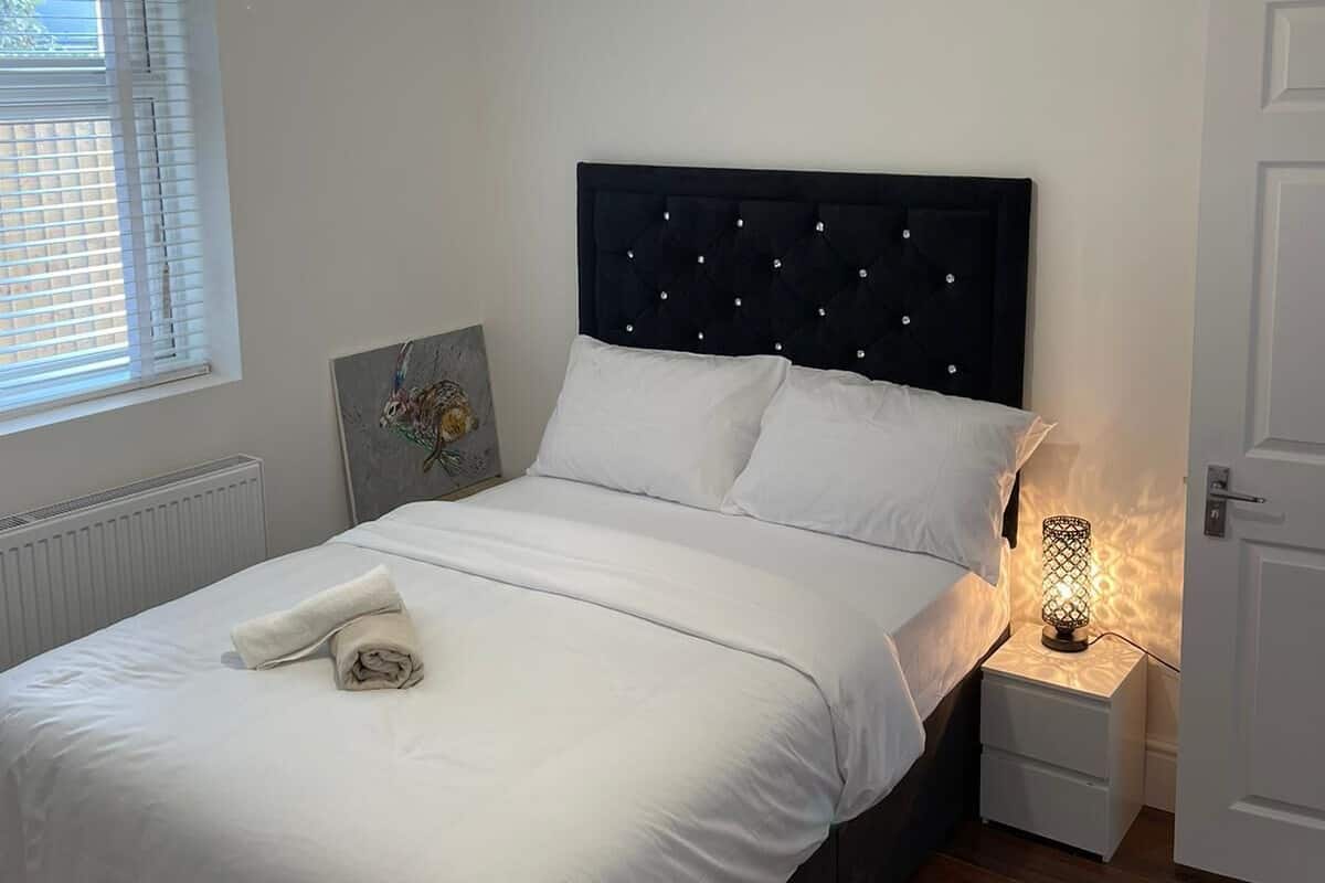 A neatly made bed with white linens, a headboard with tufting, a bedside lamp, and a radiator.