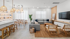 Un salón moderno con un sofá gris, sillas de madera, un televisor y una zona de cocina con mostrador y taburetes.