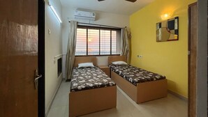 Ein Zimmer mit zwei Einzelbetten, einem Deckenventilator, einer Klimaanlage, einem Fenster mit Vorhängen und einem an der Wand befestigten Spiegel.