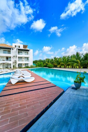 A swimming pool with lounge chairs and a brick deck, surrounded by palm trees and a modern building. - Cana Brava Residences Rental Apartment  (Punta Cana)