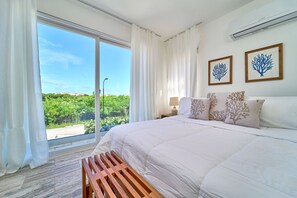 A bedroom with a large bed, white bedding, a wooden bench, a nightstand with a lamp, and two framed pictures on the wall. - Cana Brava Residences Rental Apartment  (Punta Cana)