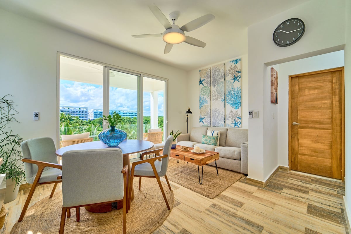 A spacious living room with a dining area, large windows, a ceiling fan, and a view of buildings outside.