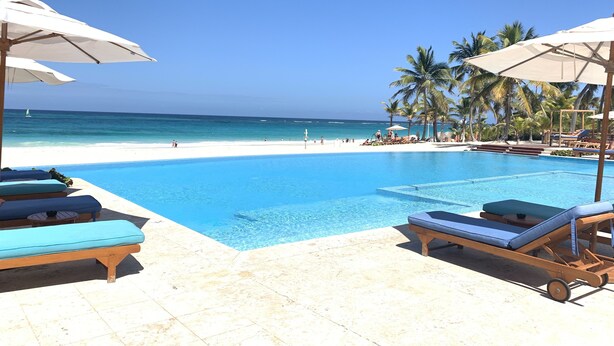 A poolside area with lounge chairs and umbrellas, a clear blue pool, and a beach with palm trees in the background. - Cana Brava Residences Rental Apartment  (Punta Cana)
