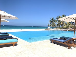 Un espace aménagé au bord de la piscine, avec des chaises longues et des parasols, une piscine à l’eau d’un bleu limpide, et une plage bordée de palmiers.